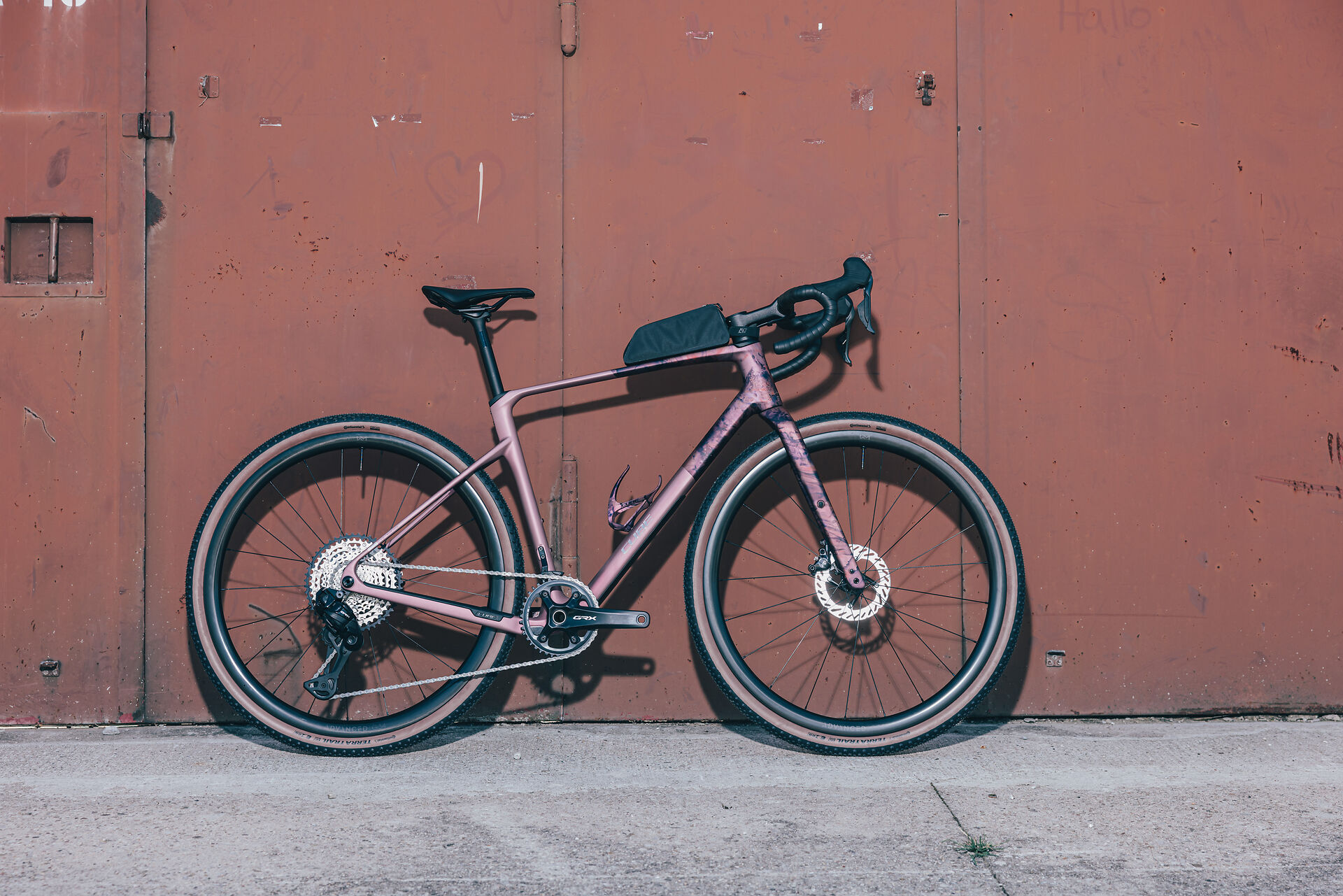 Ein rosafarbenes und lilafarbenes Schotterfahrrad mit abfallendem Lenker ist vor einer verwitterten, rotbraunen Metalltür auf einer Betonfläche abgestellt. Am Oberrohr des Fahrrads ist eine kleine schwarze Tasche befestigt.