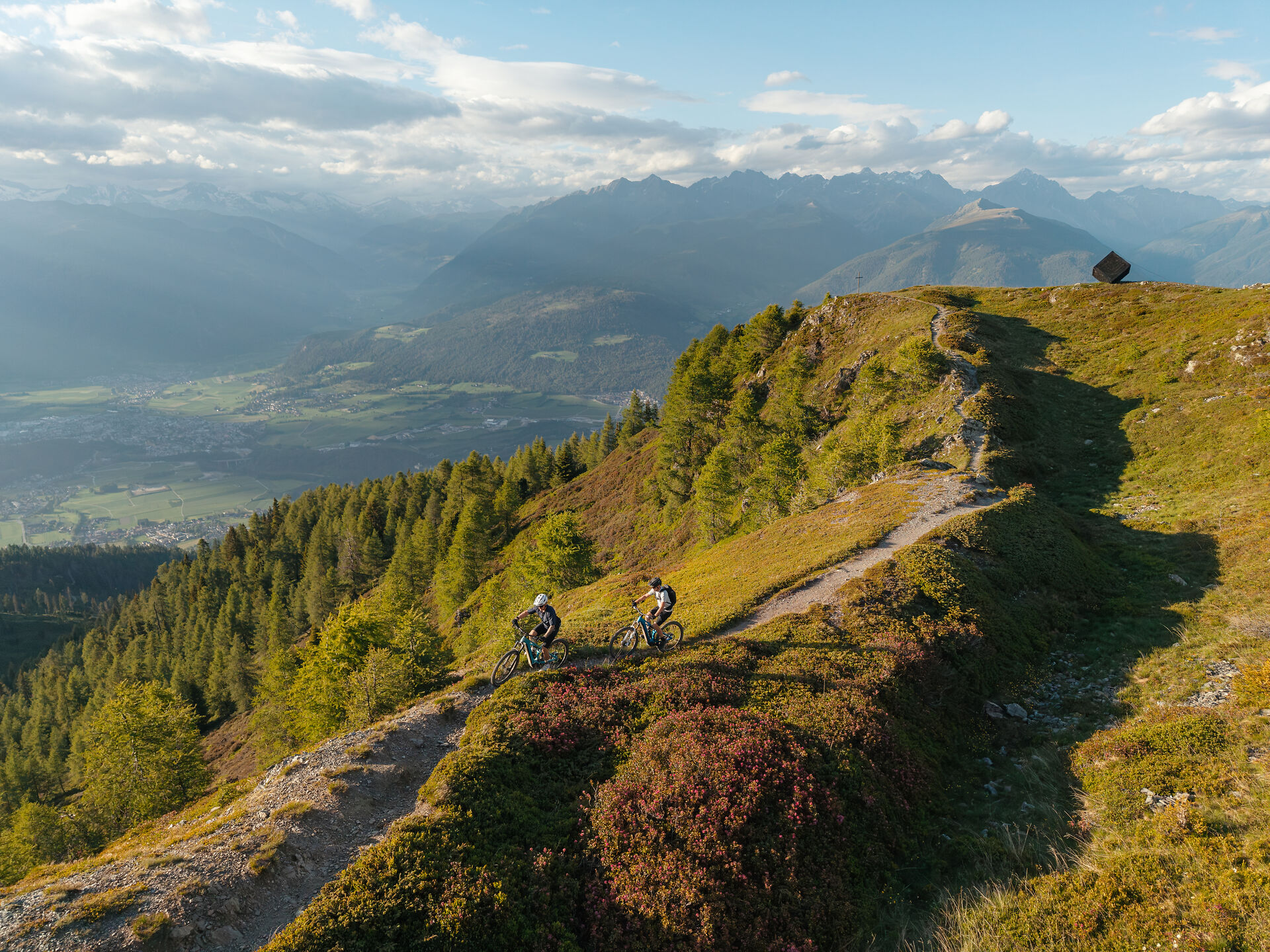 Zwei E-Mountainbiker auf einem schmalen Gratweg hoch über einem Alpental