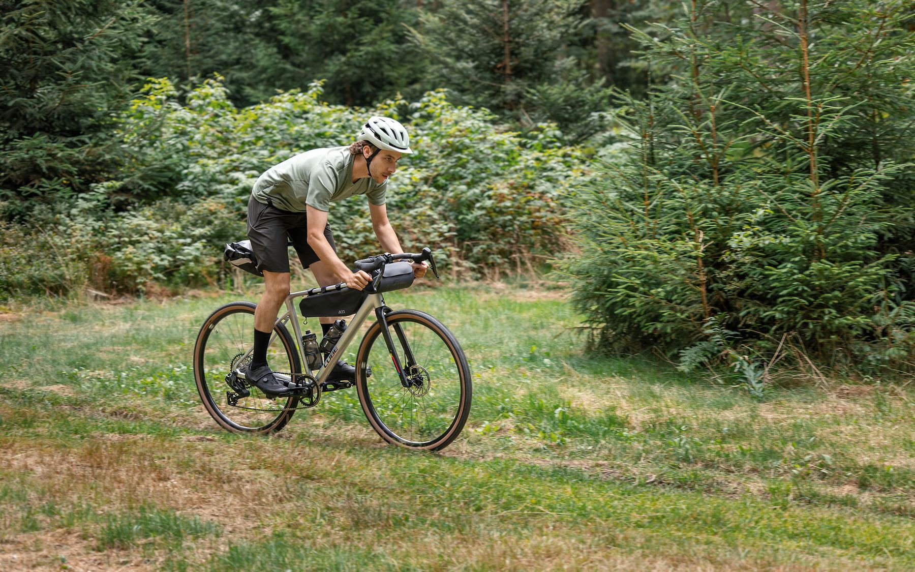 Fahrer auf einem Gravelbike mit Rahmentasche auf einem Wiesenweg am Waldrand