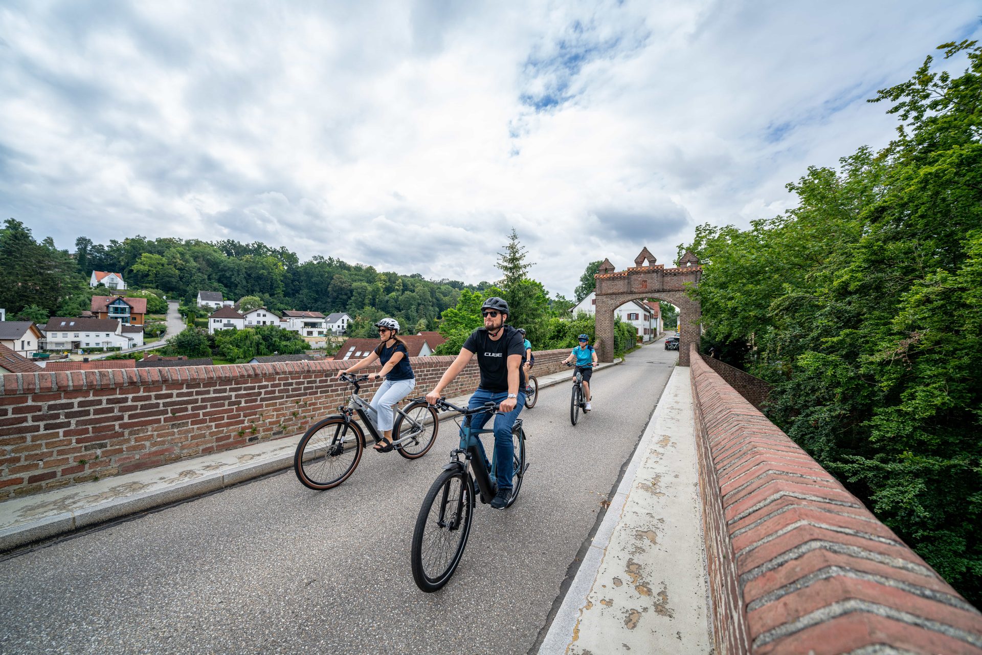 Vier Radfahrende auf E-Bikes fahren über eine Backsteinbrücke mit Torbogen in Dingolfing