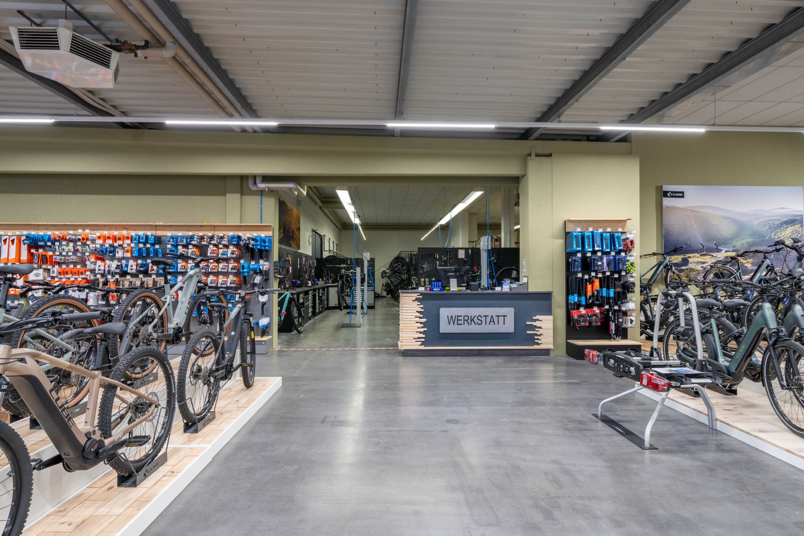 Ein modernes Fahrradladen-Interieur in Dingolfing mit Reihen von CUBE-Fahrrädern, farbenfrohen Accessoires an den Wänden und einem Servicetresen mit der Aufschrift „WERKSTATT“ in der Mitte. Das klare, übersichtliche Layout besticht durch stilvolle Industriebeleuchtung.