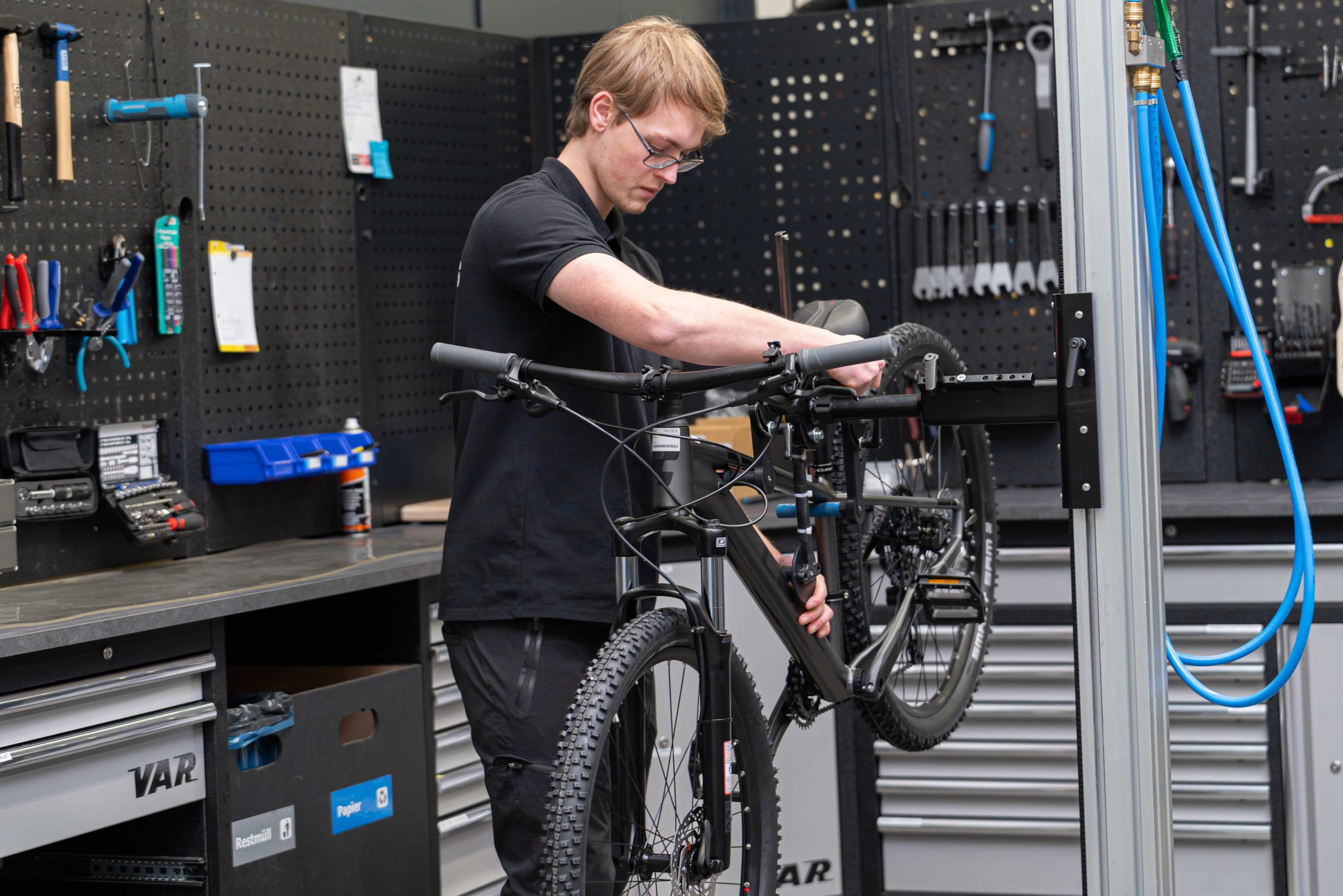 Eine Person mit Brille und schwarzer Kleidung arbeitet in einer mit Werkzeug und Ausrüstung gefüllten Werkstatt im CUBE Store Dingolfing an einem schwarzen Fahrrad, das auf einem Reparaturständer montiert ist.