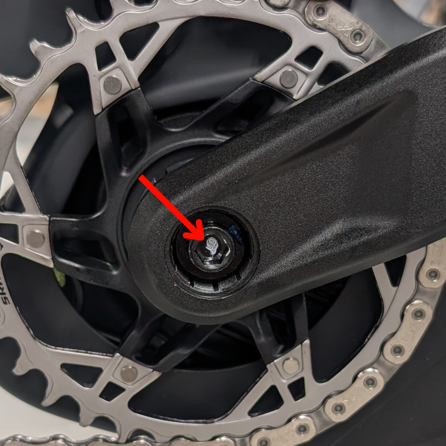 Nahaufnahme eines Fahrradkurbelarms und eines Kettenblatts mit einem roten Pfeil, der auf eine Schraube in der Mitte des Kurbelarms zeigt.