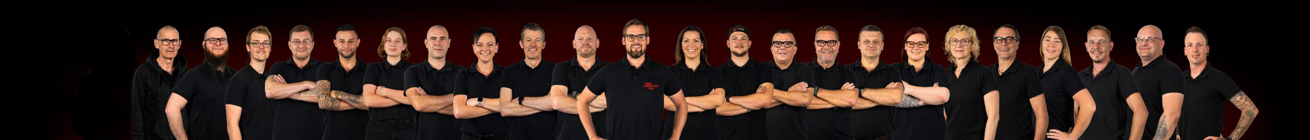 Eine Gruppe von 23 Personen steht in einer Reihe vor einem dunklen Hintergrund. Sie tragen alle schwarze Hemden und Brillen, haben die Arme verschränkt, schauen nach vorne und posieren selbstbewusst.